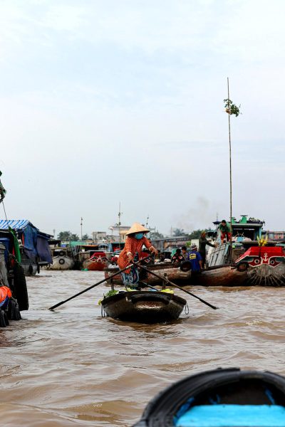 A view from the boat, on an Asian river with floating market stalls and boats in background. A woman wearing traditional Vietnamese conical hat rowing small wooden raft in brown water. There is some smoke coming out of nearby tractors driving on road above river. In distance there's tall pole with blue flag flying at top. Photo taken by Nikon d850 camera with wideangle lens, high resolution and realistic style.