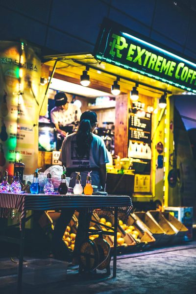 a night time photo of an outdoor coffee shop in the middle east, with neon lights and colorful decorations. A man is standing behind his stand selling drinks from a cart full of fruits and snacks. The sign above says "charger extreme Coffee". He has black hair tied back into two braids and wears streetwear . In front there's another table covered by three large open bags filled with colourful glowinthedark paint. On top one bag stands on its side with some tubes coming out of it.