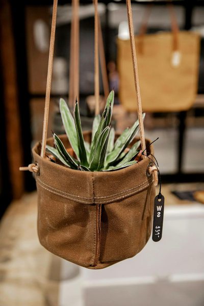 A hanging planter made from an old brown canvas bag with leather straps, displayed in the shop of W shouting Studio's brand name tag and hangin plant decor collection. The focus is on its unique design as part of their new lineup.