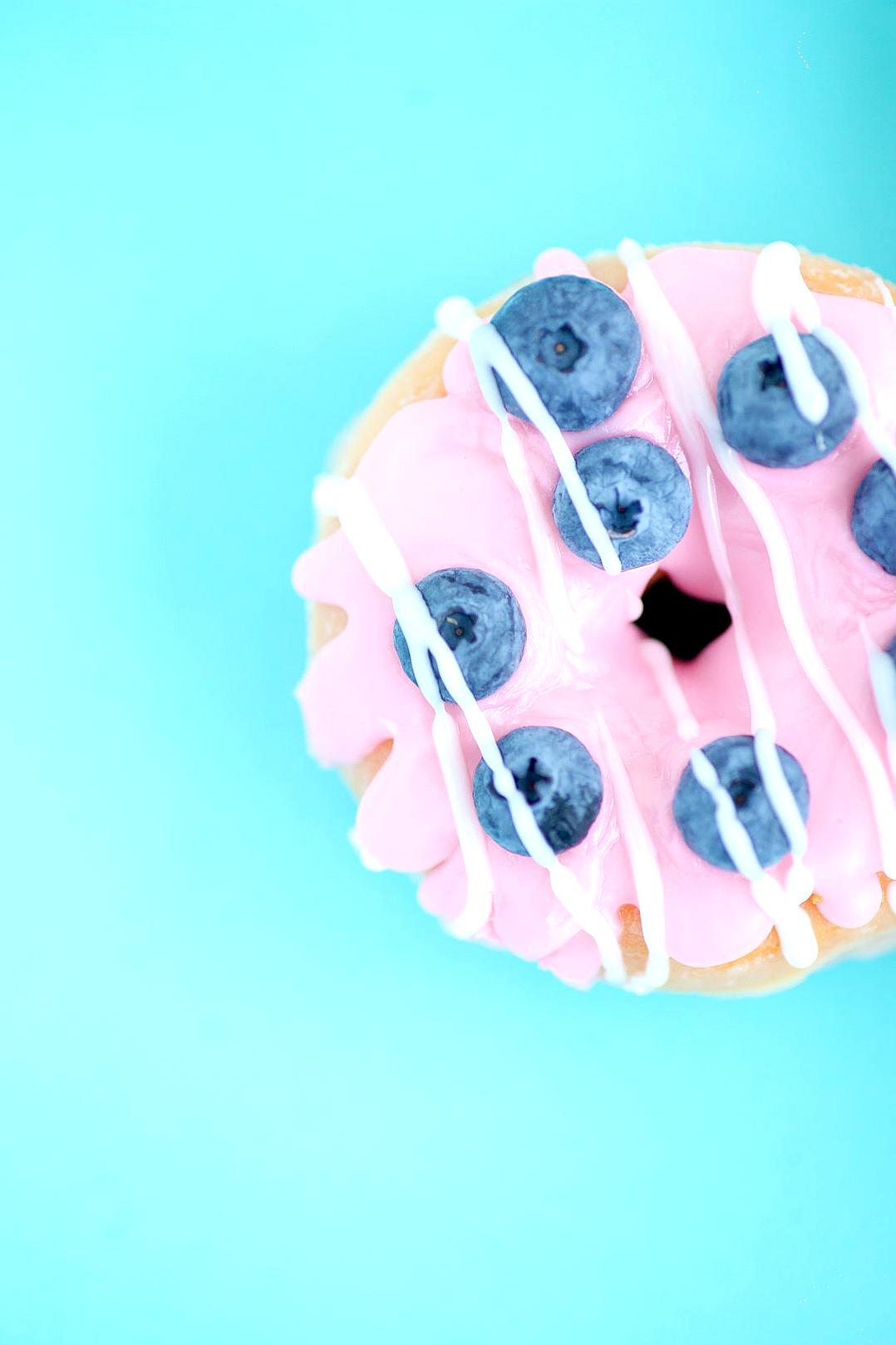 Closeup of a pink donut with blueberries and a white drizzle on top, against a solid light cyan background, in the style of minimalist photography, in a macro style, resembling food magazine photography, with high resolution