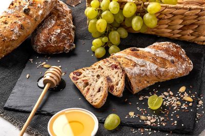 Said bread with grapes, honey and seeds on the table. On top of a black slate there is a sliced sourdough loaf. In front there's some olive oil in a small white bowl, green grapes around. A wooden spoon inside dripping a light brown glaze. The whole composition looks very appetizing. It also includes sunflower seed loaves and wheat horn shaped cookies. There's an old wicker basket behind full of fresh fruit like apples or oranges. White background.