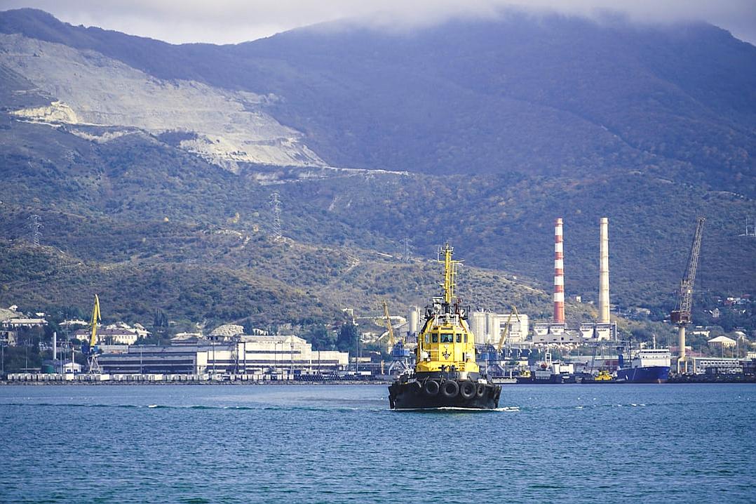 A black and yellow small tugboat is floating in the sea near Sochi, against the backdrop of mountains with an industrial complex on their shore. The cargo port stands behind it. Photo taken from water level in the style of Judge.