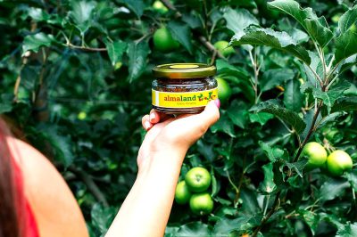 A closeup shot of the jar, held in the style of an apple orchard worker's hand with her arm extended towards camera, is captured in natural light, showcasing vibrant green apples and lush trees behind it. The focus on the jam label shows bright yellow text "alLEXandland" and brown color.