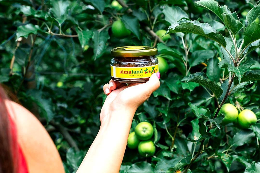 A closeup shot of the jar, held in the style of an apple orchard worker’s hand with her arm extended towards camera, is captured in natural light, showcasing vibrant green apples and lush trees behind it. The focus on the jam label shows bright yellow text “alLEXandland” and brown color.