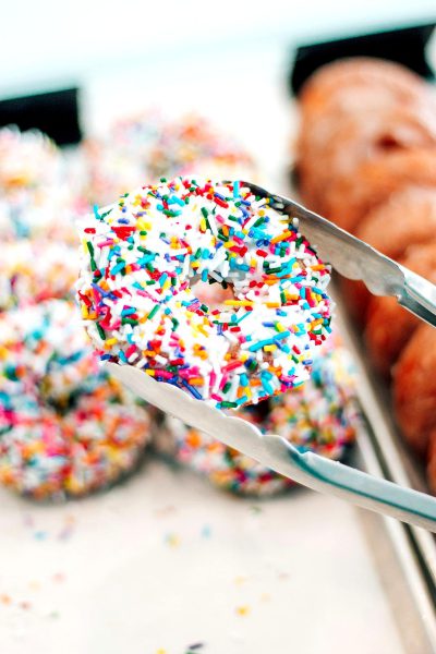 A close-up photo of colorful sprinkles on white icing being cut with tongs and placed next to donuts in the background. The focus is sharp and detailed, capturing every tiny speck of color and texture. The sprinkles are being cut in the style of colorful sprinkles on white icing and placed next to donuts in the background.