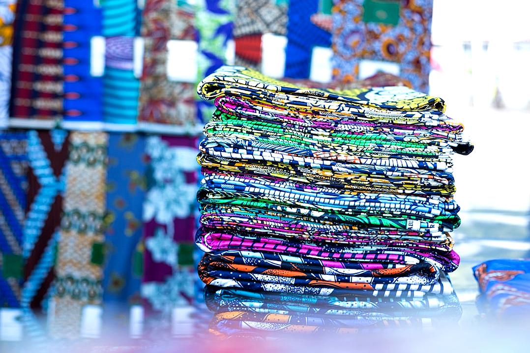 A stack of colorful African fabric was displayed on an urban street market stall, with various patterns and designs in the background. The fabrics appeared vibrant against the cityscape backdrop, capturing the essence of traditional fashion and cultural heritage in Africa. Shot in the style of Canon EOS R5 camera using natural light, the focus was sharp to highlight intricate details within each pattern.