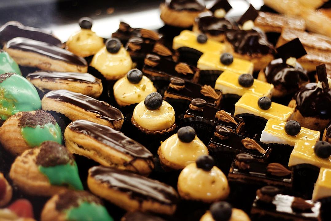 A closeup of an assortment of French pastries, including chocolate-covered eclairs and green-tinted crVoisins, arranged on display at the bakery’s counter. The focus is on intricate details like shiny black chocolate lines or glossy yellow cream filling in each pastry. This scene captures the vibrant colors and textures that make these mini confections one-of-a-kind pieces of artistry in the style of French pastry artists.