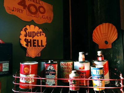 A vintage gas station display featuring "The Super shell dry fuel" sign, an orange seashell logo, and various antique oil cans on the wall. A wire rack holds old advertising from The golden age of car photography.