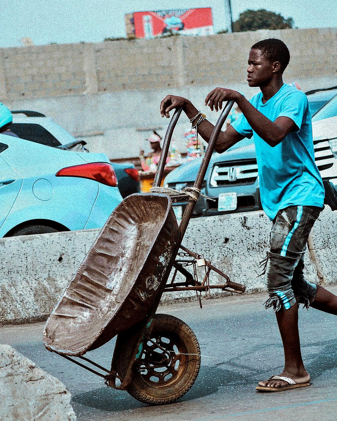 photograph of an african young man wearing blue t shirt and short pants, pushing wheelbarrow full with car parts in the parking lot, he is holding his wrist on top of it