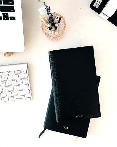 flat lay of a black leather notebook on a white desk with minimalist office accessories. The cover has "Kypical" written in gold lettering, in the style of minimalistic style. Natural light product photography.