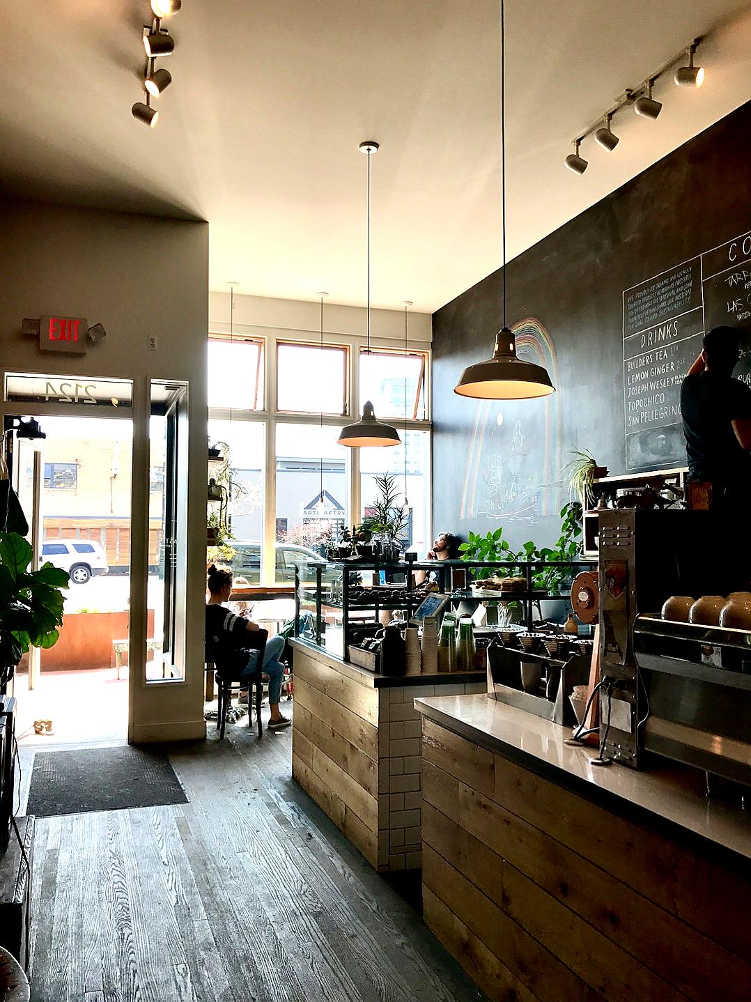 A photo shows the inside and entrance of an urban coffee shop with wood accents. A chalkboard menu hangs on the wall, with hanging pendant lights above the counter top. In the background, a large window looks out onto the street in downtown Port arena BC Canada. The vibe feels cool yet welcoming, modern and rustic. There is one person sitting at a table drinking a latte. It is daytime and natural light comes through the windows.