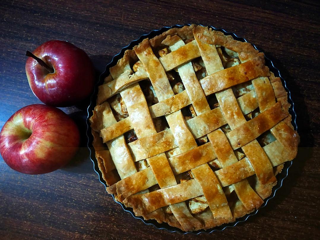 A large apple pie with a latticed crust, sitting on top of an old wooden table next to two red apples. The whole scene is captured in a photo taken from above.