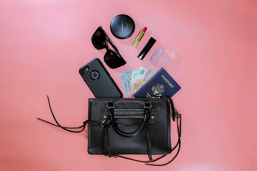 A black leather handbag lying on the floor with items spilling out of it, such as sunglasses and lipstick, next to an iPhone in its case, A passport laying flat against pink background, high resolution