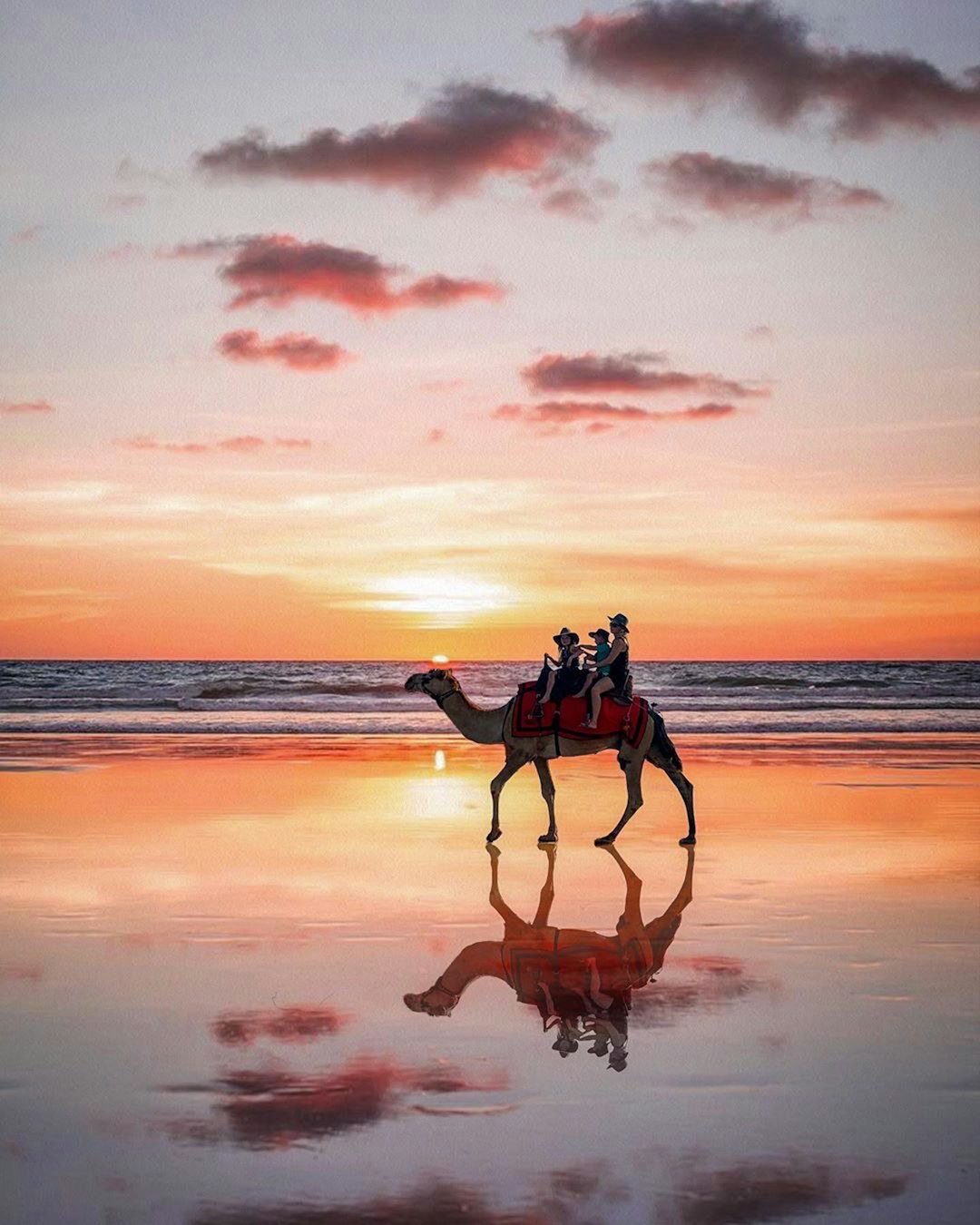 A photo of two people riding on the back of an animal at sunset, vibrant orange and pink sky reflecting in water with one person wearing red sitting atop camel as it walks along a flat white sand beach, in the style of cinematic. –ar 51:64