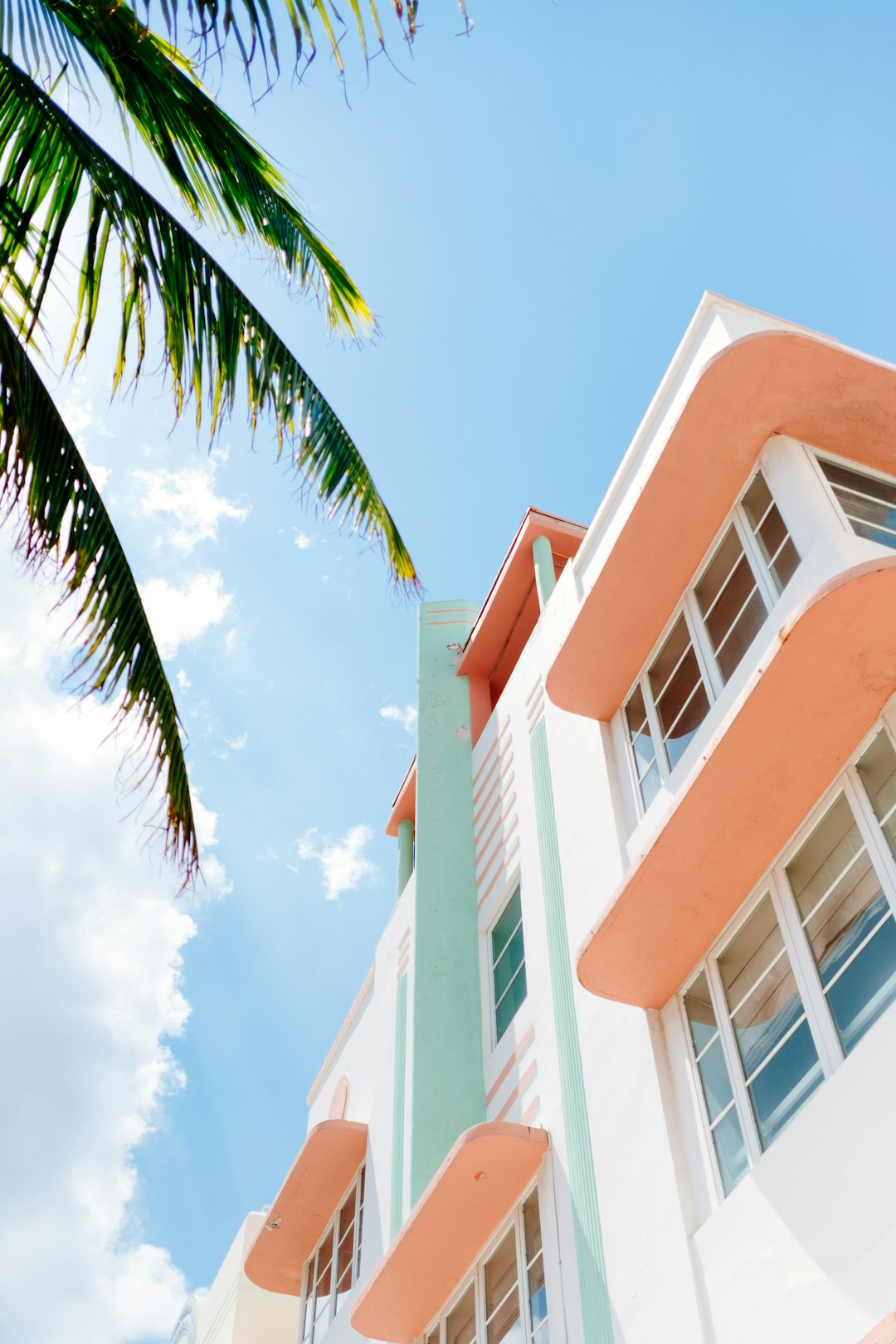 miami art deco buildings, white walls with pastel orange and pink accents, palm tree, blue sky, perspective, low angle, editorial photography, –ar 85:128