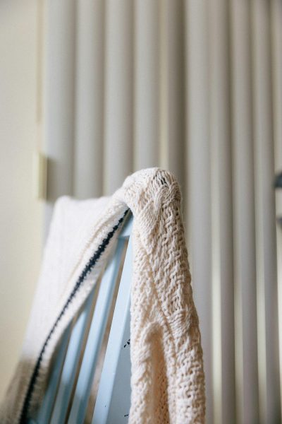 A closeup of the edge of an ivory knit sweater draped over the armrest of a baby blue rocking chair, with vertical white blinds softly focused behind it. The sweater's texture is highlighted by natural light, and its black trim adds contrast to the room’s neutral color scheme. This shot emphasizes the cozy feel created through the combination of fabric and wood in the style of a natural light photographer.