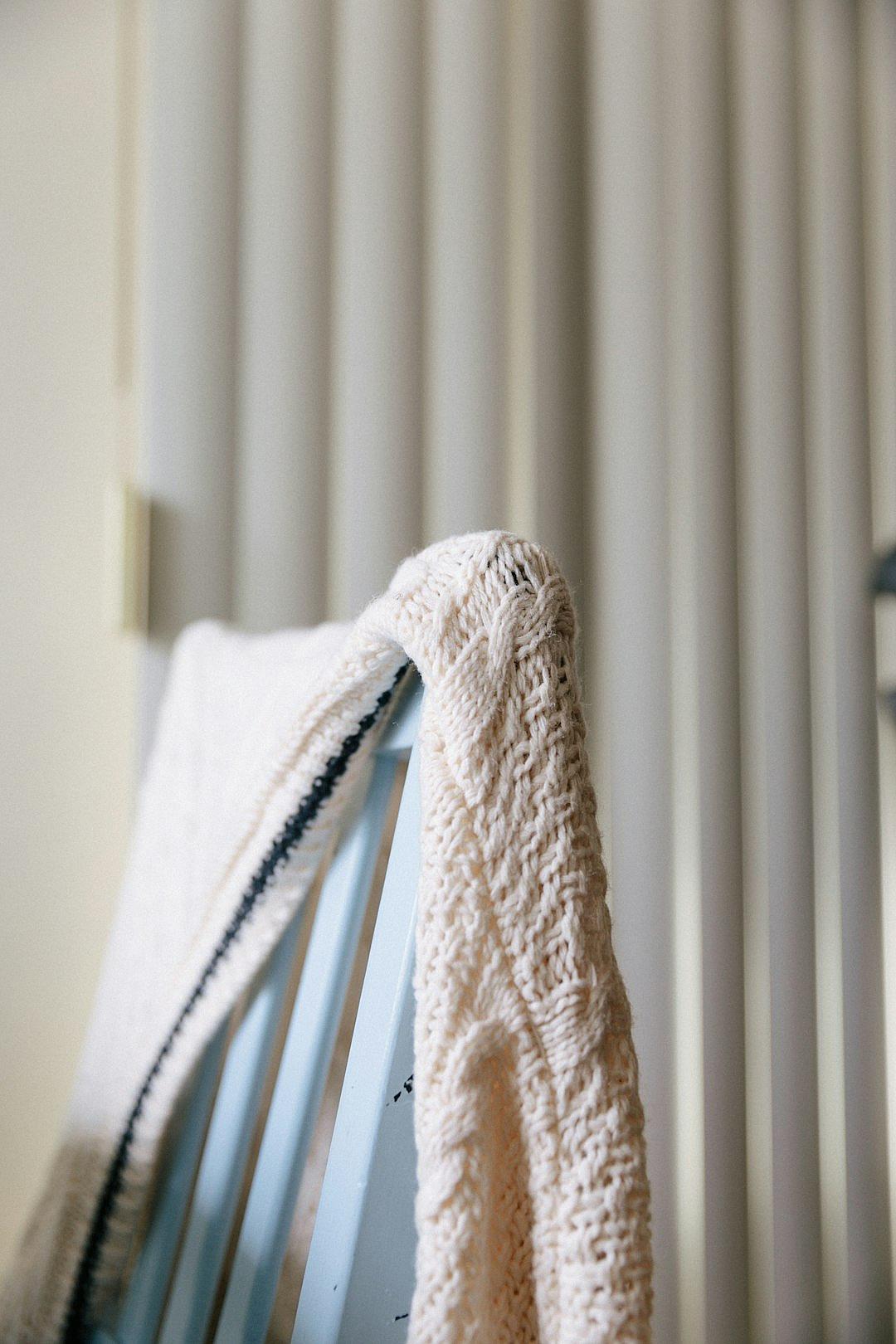 A closeup of the edge of an ivory knit sweater draped over the armrest of a baby blue rocking chair, with vertical white blinds softly focused behind it. The sweater’s texture is highlighted by natural light, and its black trim adds contrast to the room’s neutral color scheme. This shot emphasizes the cozy feel created through the combination of fabric and wood in the style of a natural light photographer.