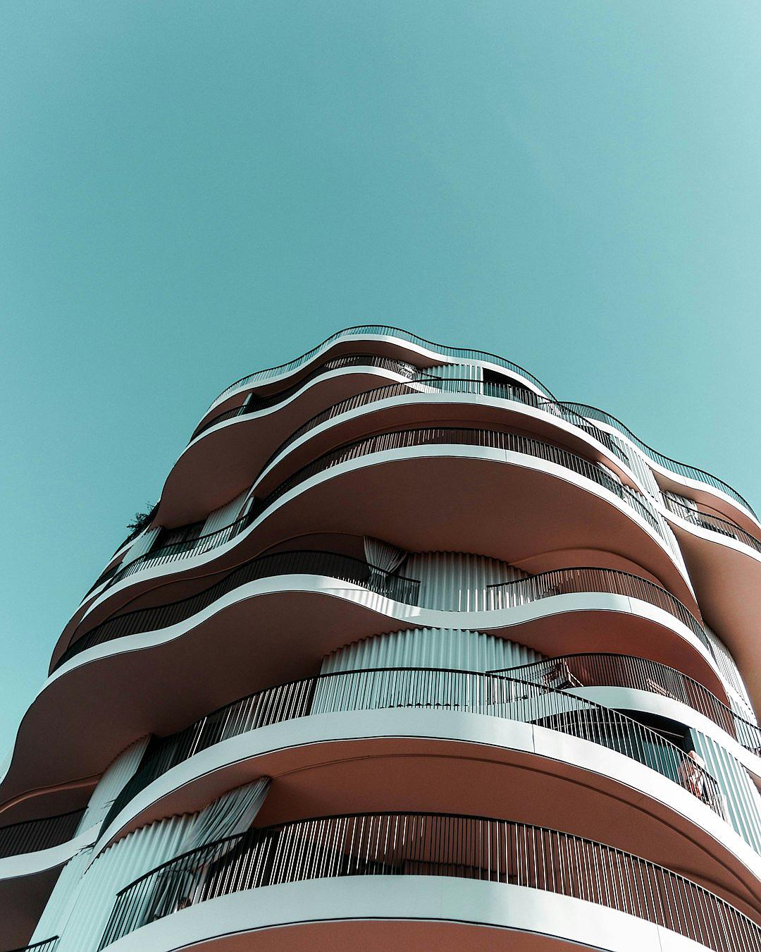 low angle photo of a curved residential building with balconies, peach colored facade, minimalist design in the style of [Bjarke Ingels](https://goo.gl/search?artist%20Bjarke%20Ingels) against the blue sky