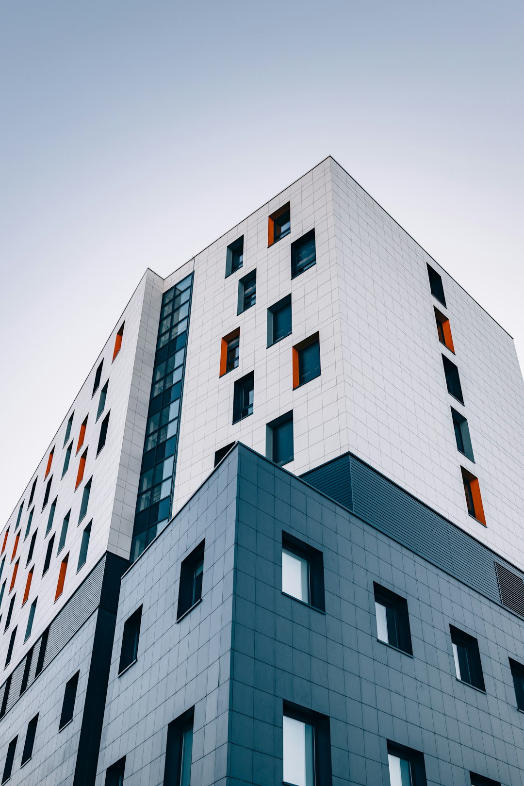 A modern building with white and grey tile walls, orange windows, and blue accents. The sky is clear in the background. A low-angle shot capturing the sharp angles of its exterior design. Shot on Canon EOS R5 mirrorless camera using an aperture setting of f/2.0 to create shallow depth of field. High resolution. –ar 85:128