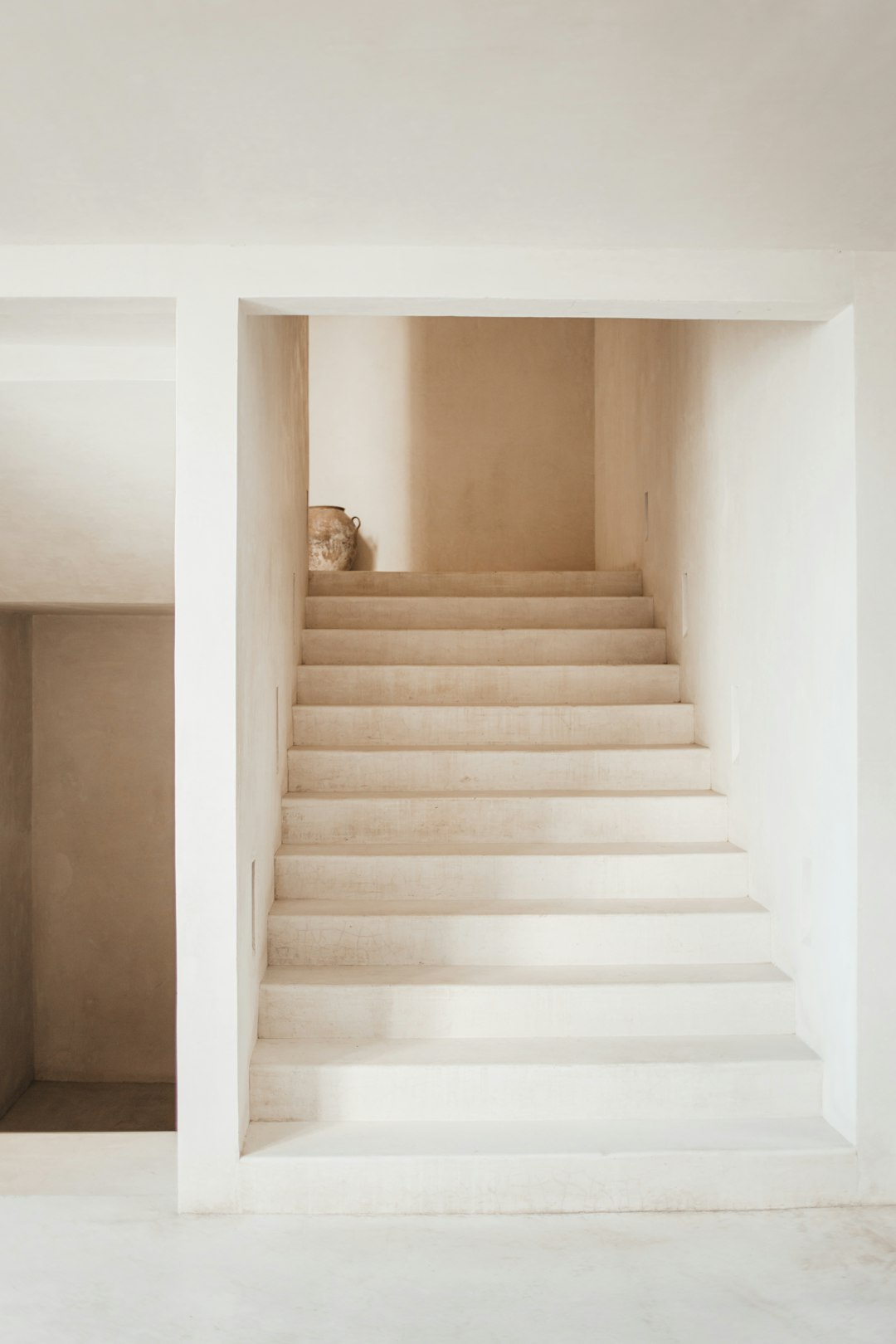 A staircase leading to the upper floor of an apartment, with smooth white walls and concrete stairs. The space is simple yet elegant, featuring subtle details like a small vase or ceramic figure on one side for decorative contrast. This photo was taken by Alvaro Paris using Canon EOS camera and natural light, showcasing the minimalistic design in neutral tones. –ar 85:128