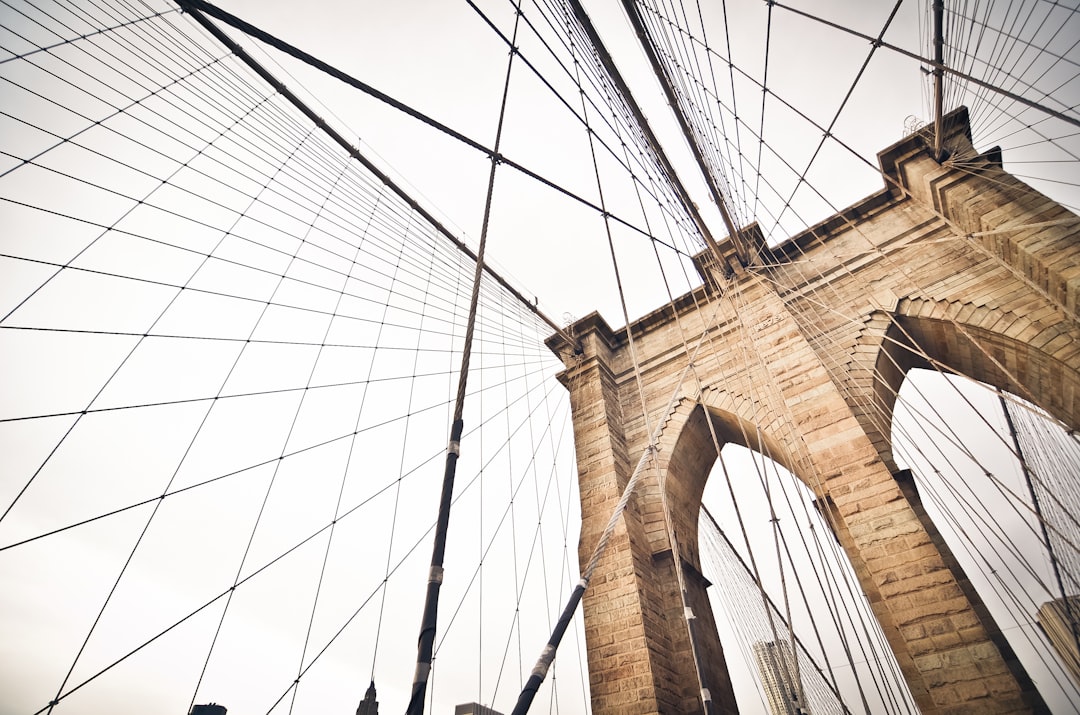 A close-up shot of the Brooklyn Bridge, showcasing its intricate architecture and unique architectural elements. The photo captures the detailed textures on the stone arches and cables against a white sky background. This perspective adds depth to the scene, highlighting one iconic landmark in New York City. Shot with a Canon EOS camera using natural light and a wide-angle lens for a clean composition. High resolution.