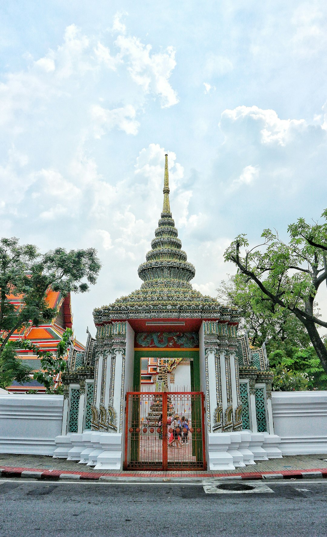 A photo of the entrance to Phra Chetrapavesa in Bangkok, Thailand with traditional Thai patterns and colors on its gate, set against an urban backdrop. The temple is decorated with vibrant green and red designs, surrounded by white walls and trees under a cloudy sky. This scene captures nature’s beauty contrasted with architectural elegance, shot from the front angle.