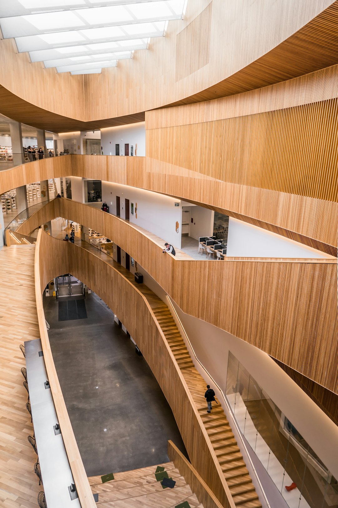 Aerial view of the interior design of a layered wood and glass building with curved wooden staircases in an open space environment with people walking around. The office building was designed in the style of [Bjarke Ingels](https://goo.gl/search?artist%20Bjarke%20Ingels) and is located on a river bank surrounded by a boxwood forest. Natural light and soft shadows are seen in the interior architecture photography. –ar 85:128