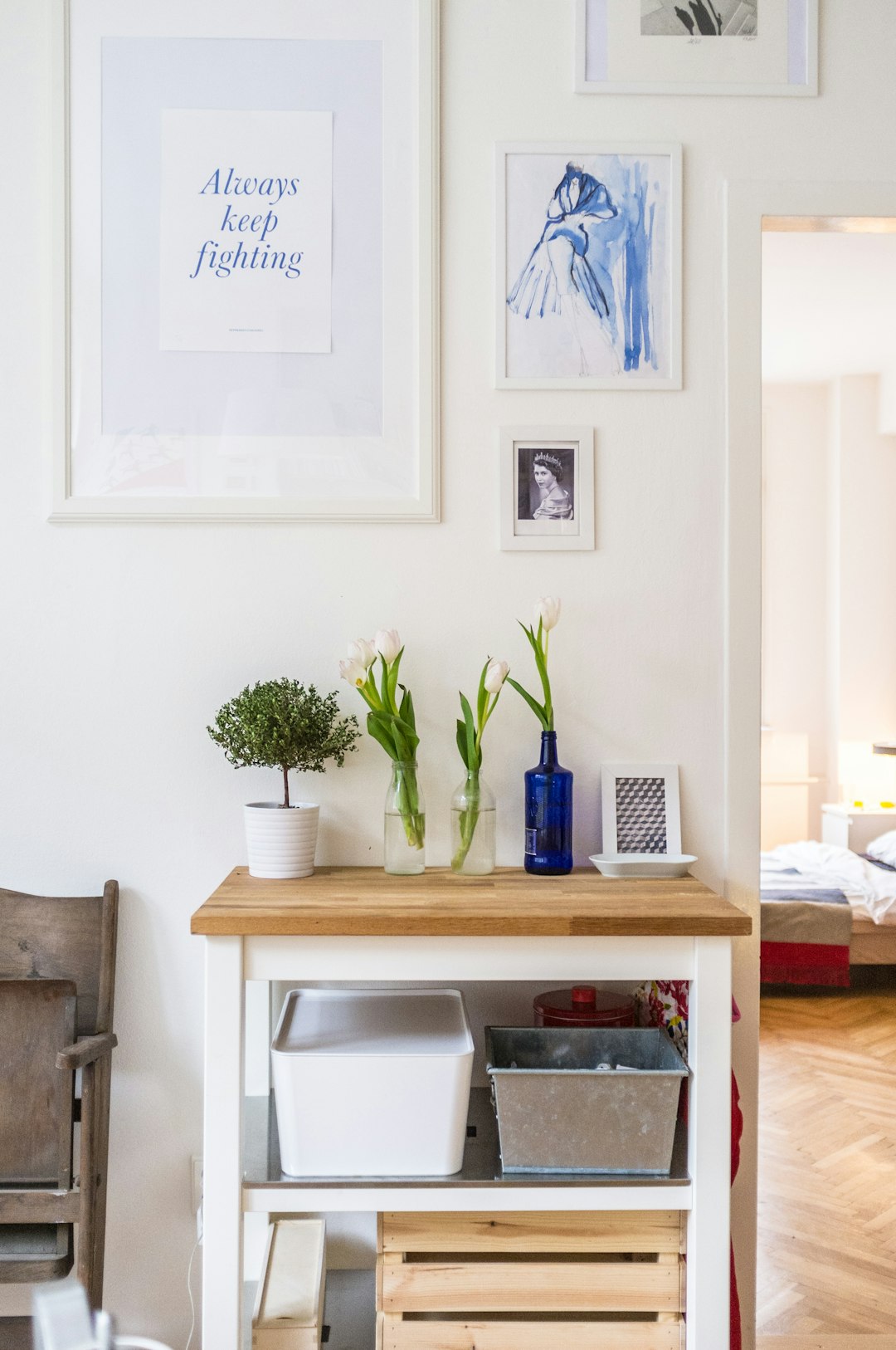 A Scandinavianstyle interior design of an apartment, featuring white walls and wooden floors. A small table with wood top is placed in the center on which there’s some storage boxes, vases holding flowers like tulips or blue roses, photo frames, books, blue glass bottle with cap. On one wall hangs three framed posters with text “bbie always keep fighting”. The overall mood exudes simplicity, clean lines, natural light, tranquility, minimalism, and warmth.