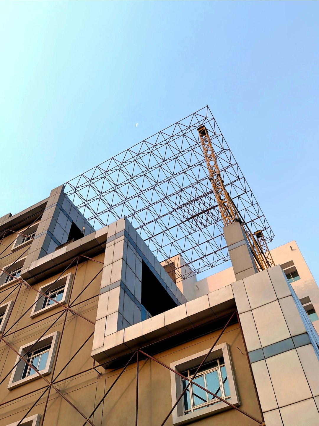 A photo shows an outdoor construction site in New Delhi with a flat tiled area. A modernist building is being built on the top floor. The sky is a clear blue and sunny. A large metal grid structure stands tall above it. It is made up of interlocking triangular elements that form its main frame, creating a geometric patterned appearance. There is no roof or ceiling visible yet, making for a striking contrast against the bright daylight. One crane can be seen working nearby.