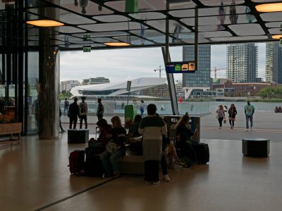 A photo of the interior of Central Station in the detectorship area, people sitting on chairs with luggage and waiting for their train, modern architecture, the building is made of glass and steel, outside there is an open space overlooking the river IJ with the Amsterdam skyline behind it, the sky has some clouds, in the background you can see a big white concert hall, in the style of a photo taken with a Canon EOS R 50mm f/8s lens.