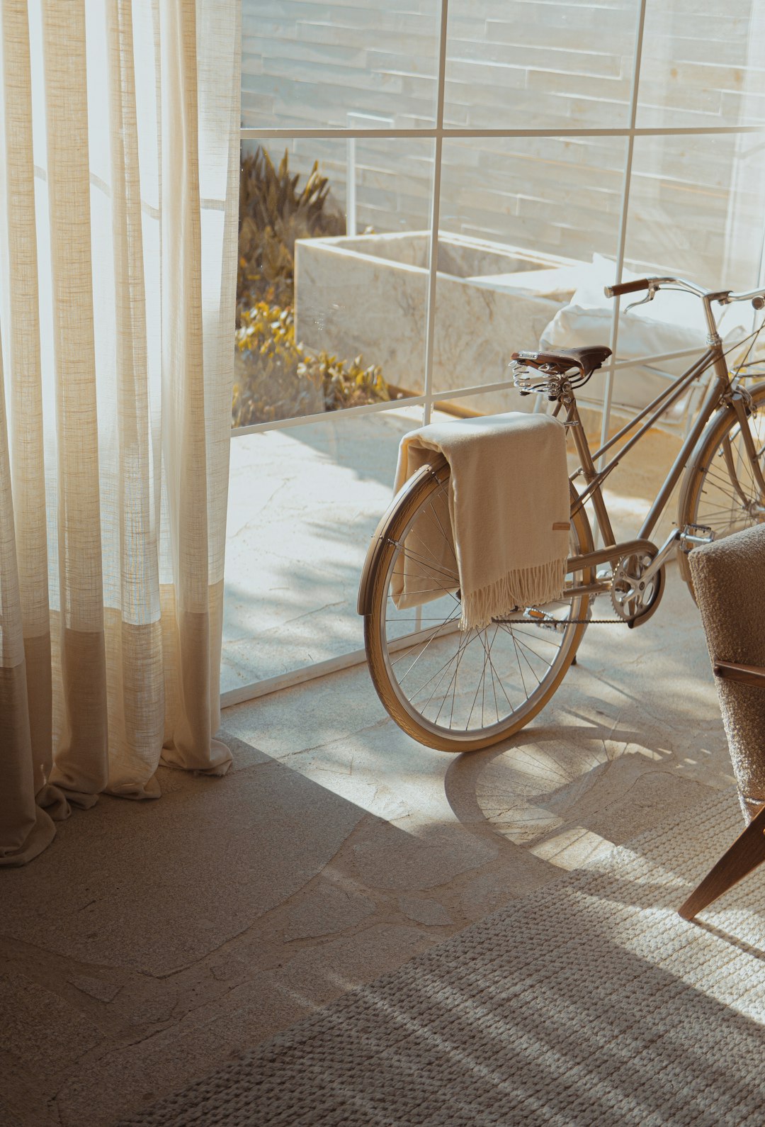 A bicycle is parked in the living room, with white curtains and sunlight shining through them on the floor next to it. The background features an outdoor swimming pool. A beige blanket hangs from one of its wheels, creating a warm atmosphere. Minimalist composition style, light gray tones, and a sense of luxury. Shot in the style of Canon EOS R5 F2 ISO30 89mm f/4. –ar 87:128