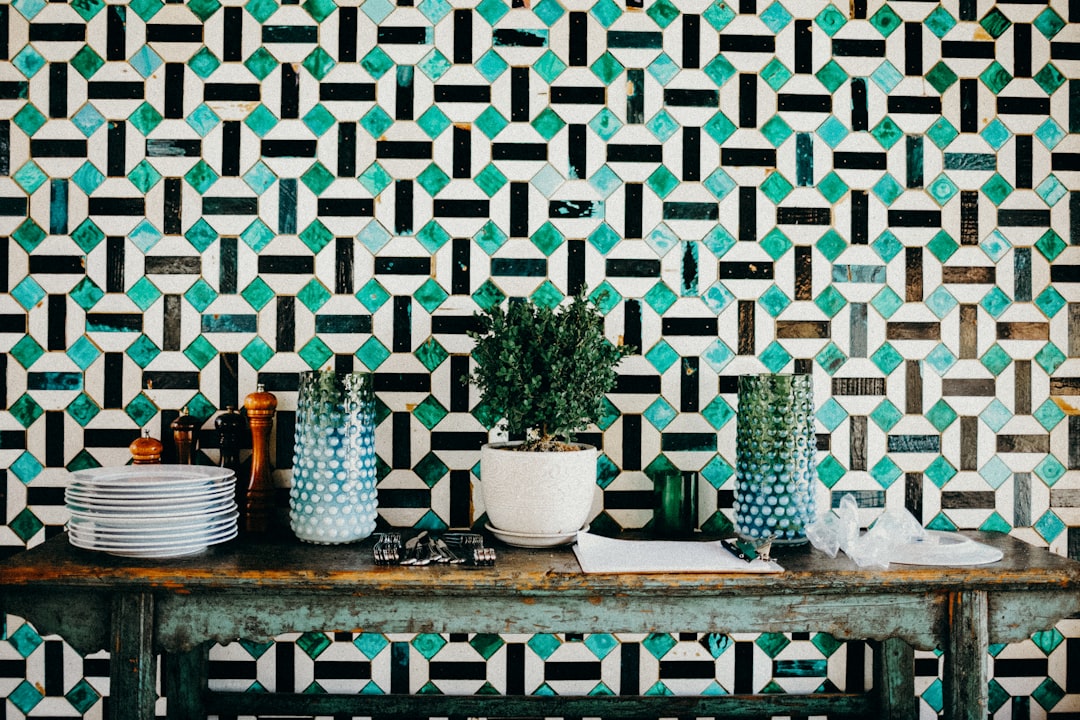 A dining table with green and white patterned tiles, geometric patterns in black and teal, potted plants on the side of an old wooden console, plates and glasses stacked on top, vintage kitchen style, bohemian inspired wallpaper. –ar 128:85