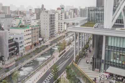 A view of the street from an elevated platform in Tokyo. There is a river running through buildings and people walking along it. There is green space on a roof with a seating area. Modern architecture and people dining outside a restaurant nearby. A grey sky, white walls, and white lines crossing the streets on the ground. A wide angle shot in the style of modern architecture.