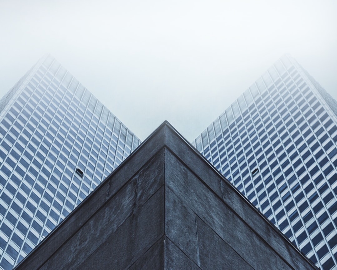 low angle photo of two skyscrapers, foggy day, grey sky, building corner, architectural photography, minimalist. The photo shows two skyscrapers on a foggy day with a grey sky taken from a low angle at the corner of a building in the style of minimalist architectural photography.