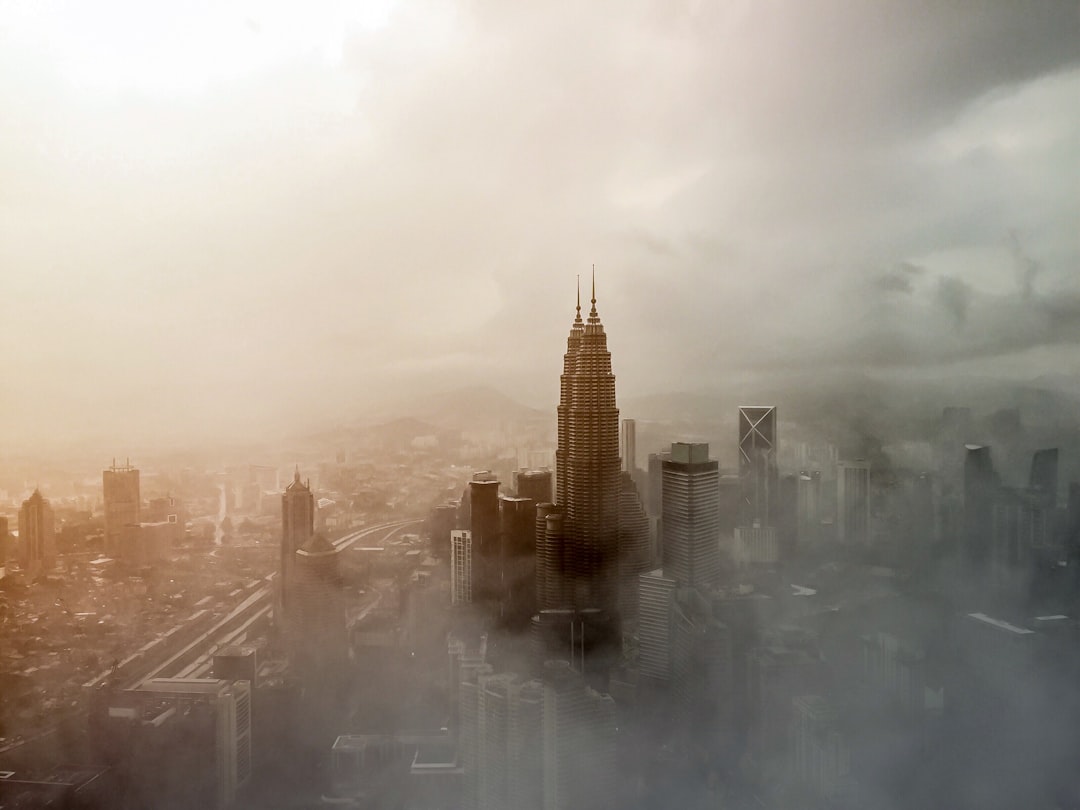 Cinematic shot of the Petronas Tower in hypnotic fog, surreal, hyper realistic, ultra high resolution, golden hour, soft light, hazy, city skyline, distant mountains, overcast sky, motion blur, grey haze, hilly landscape, tall buildings, misty clouds, grey skies, overcast day, foggy atmosphere, smoky, distant hills, grey skyscrapers, cityscape, urban environment, overcast lighting, grey clouds, distant hills, distant mountain range, distant sky. –ar 4:3