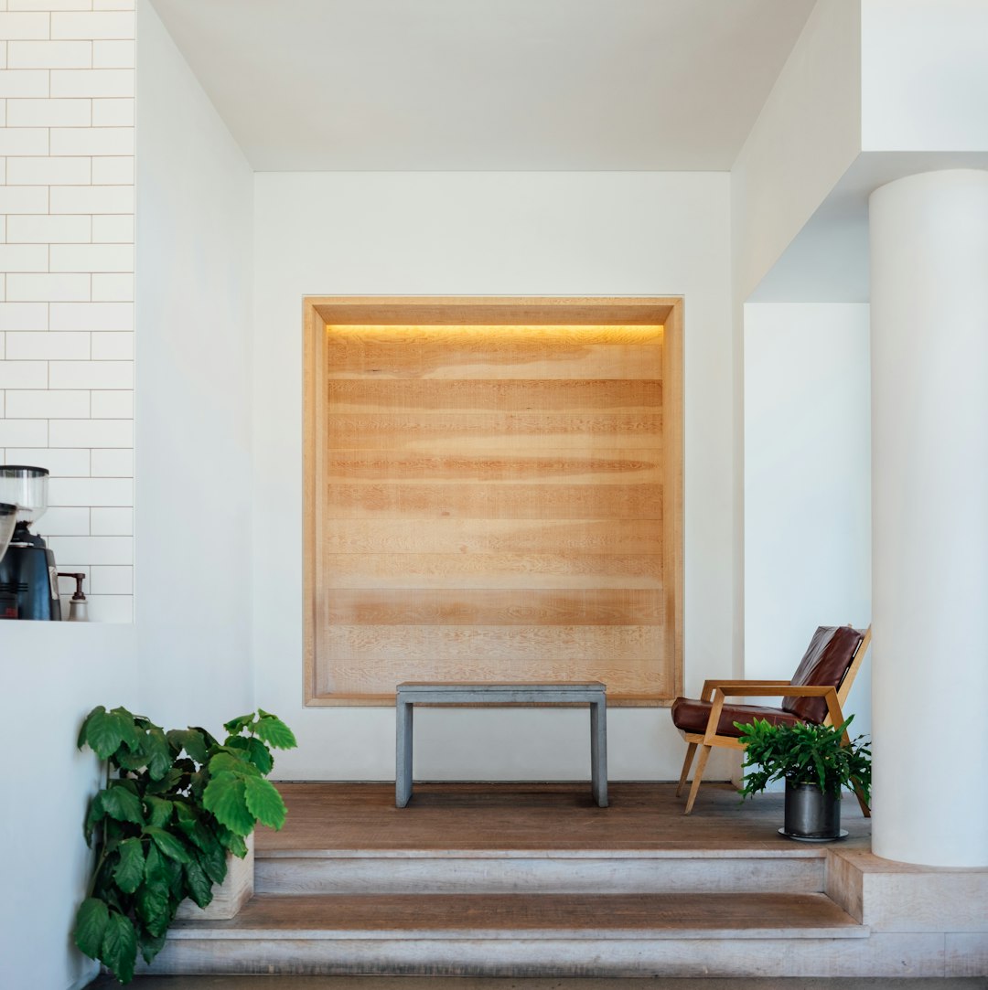 A wooden frame with light strips inside on the wall of an empty white room, with wood flooring and stairs leading to another level. There is also a coffee table and chair in front of that. The photo was taken from behind and above the entrance area, with plants placed next to it. This picture has been created in the style of [James Turrell](https://goo.gl/search?artist%20James%20Turrell). It features a modern minimalist style with soft lighting.