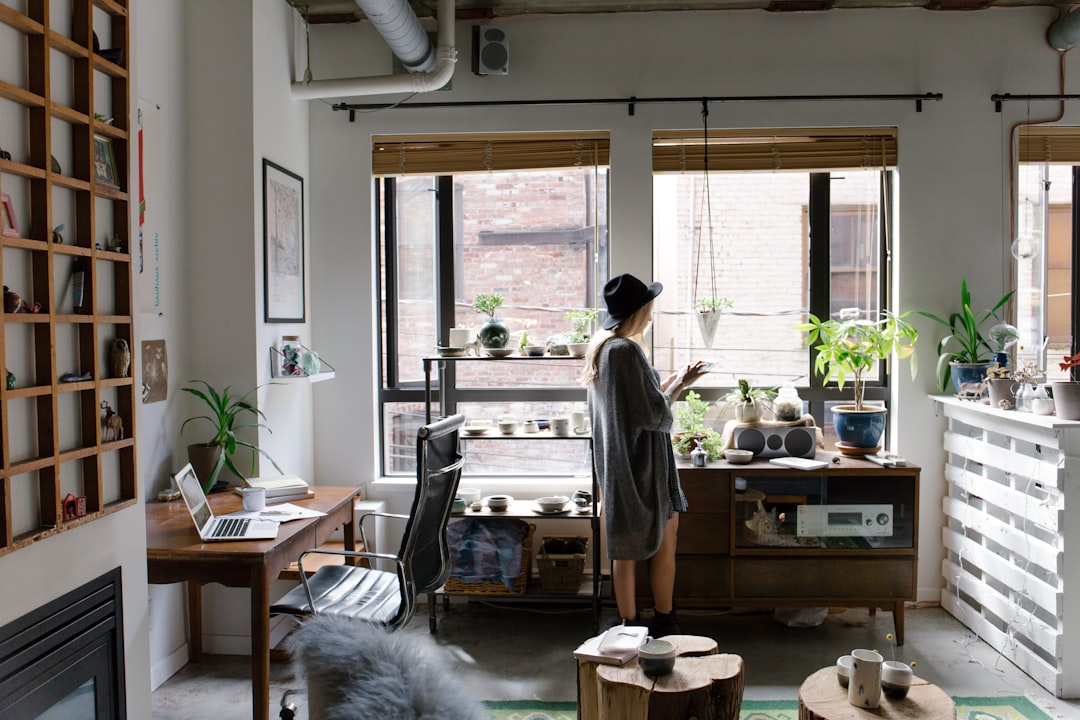 A photo of an urban loft apartment with large windows, with natural light streaming in. A woman stands near the window, wearing comfortable  and a hat. She’s reaching for plants on shelves or holding coffee mugs while standing next to her desk with tech gadgets and decor items. The room is decorated with wooden furniture, potted greenery, modern art pieces, and vintage accents, creating a cozy yet stylish space that reflects its edgy city vibe. The photo is in the style of an urban artist known for capturing loft living spaces in a natural yet stylish manner.