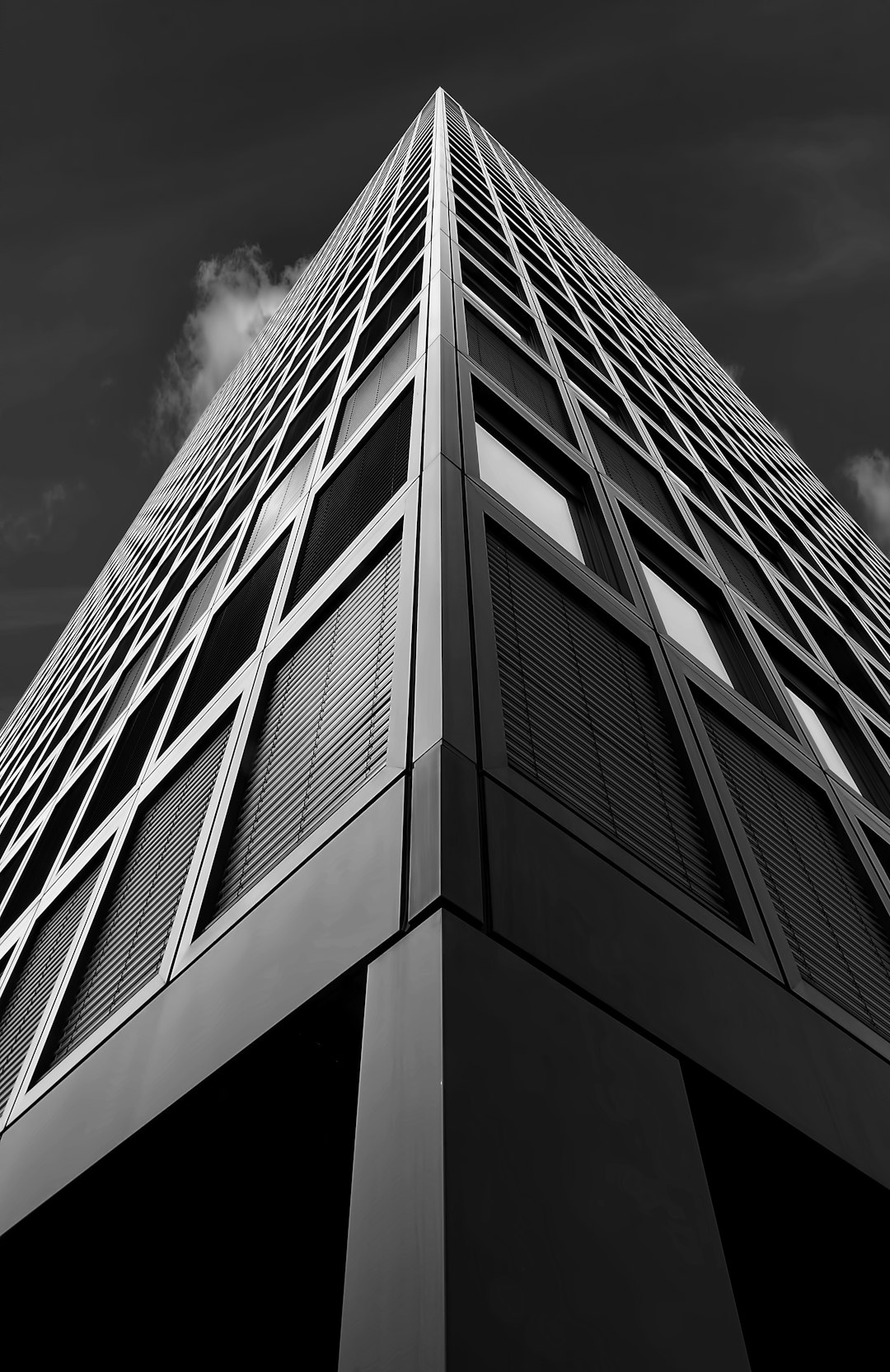 A tall building with triangular windows, captured from the ground looking up at its sharp corner in black and white. The sky is dark above it, creating contrast between light and shadow on the building’s surface. This photo was taken in the style of photographer Peter Haw, a photography award winner. It showcases the architectural design of that skyscraper. Shot with a Sony Alpha A7 III camera using an f/8 aperture setting to capture details and depth of field. –ar 83:128