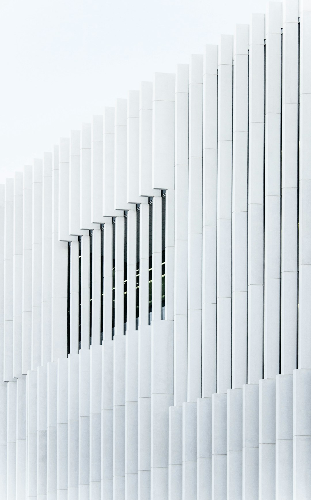 A minimalist white building facade with vertical slats that create an illusion of depth, photographed from the side against a pure background. The slabs form sharp angles and display intricate patterns in their texture, adding visual interest to the composition. –ar 79:128