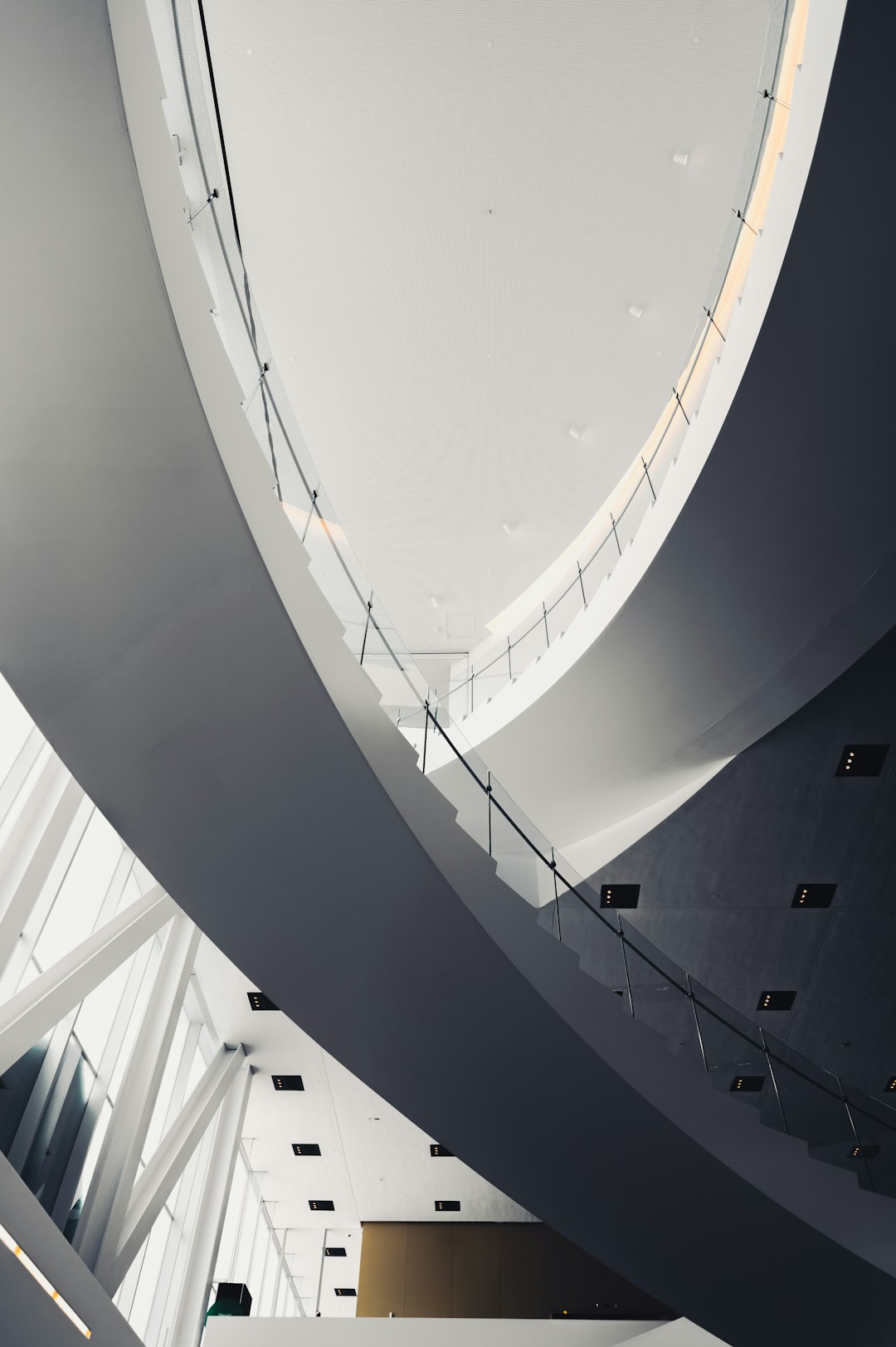 A view of the ceiling and balcony in an art museum designed in the style of [Bjarke Ingels](https://goo.gl/search?artist%20Bjarke%20Ingels) with glass balustrades, curved white steel beams, and a dark grey metal structure. The architecture is modernist with clean lines and minimalist design elements. There is no furniture or people visible inside, creating a sense of solitude and contemplation within the space.