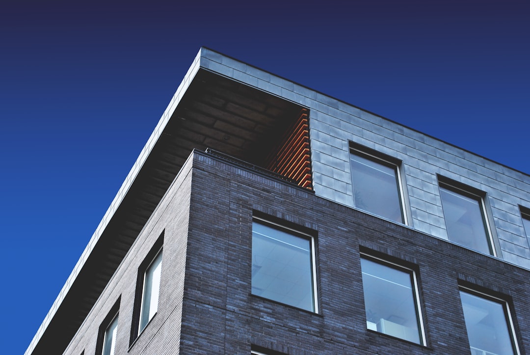 A close up photo of the corner exterior wall of an apartment building made from dark grey brick with large windows, a flat roof and an angle on one side showing blue sky. There is some wood cladding on top. The focus point should be at eye level so you can see all three floors of the building. The shot was taken in the style of Sony Alpha A7 III camera with a wideangle macro lens at f/8 aperture for a shallow depth of field effect. –ar 128:85