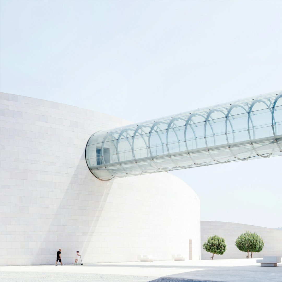 A white museum building with an arched glass bridge in the middle, in a minimalist style, on a pure sky blue background, minimalist landscape photography in the style of architectural photography, a curved shape, white walls and tiles on one side of buildings, two people walking around, minimalism in the style of architectural photography, minimalist architecture in the style of architectural appearance, architectural details, archdaily architectural photographer.