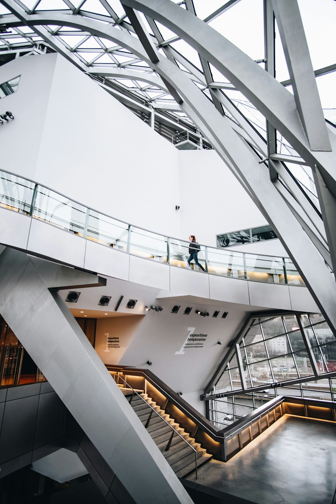 A modern museum interior with a steel frame architecture, white walls and glass windows. A person is walking on the top floor of an office building in the center. The stairs lead down to another level below. There is some natural light coming through from outside. Captured using a Canon EOS R5 camera with a standard lens, in the style of no particular artist.