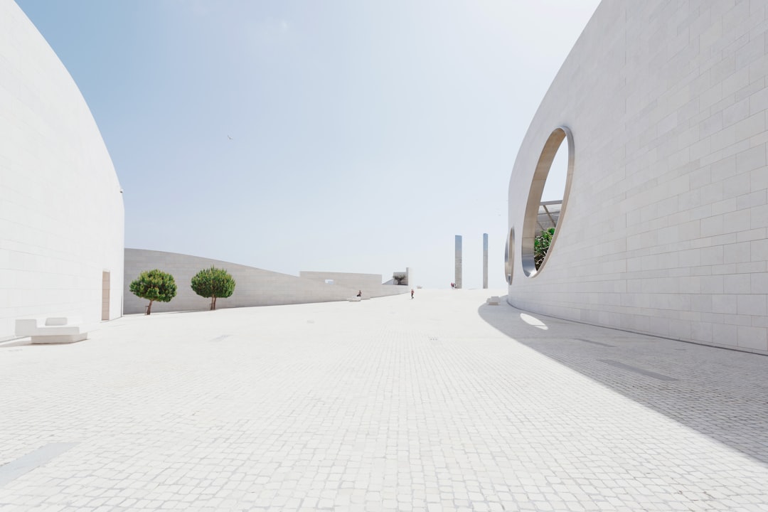 A wide-angle view of the outdoor courtyard at Al He.Fields in Doha, editorial photography, pure white walls with circular windows, minimalism, minimalist landscape design, curved lines and shapes inspired in the style of Greek architecture, stone pavement, trees, plants, blue sky, sunny day, clean and simple, architectural masterpiece, award-winning photography, architectural magazine. –ar 128:85