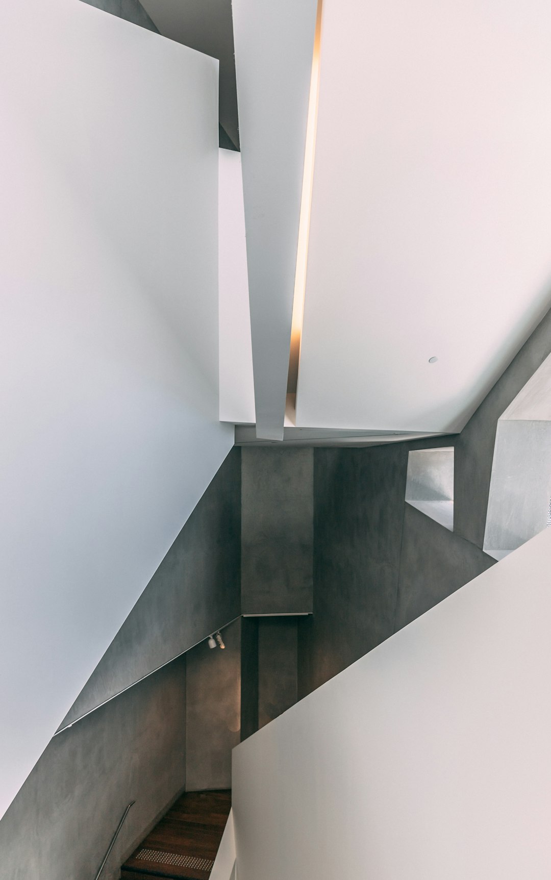 A staircase in the interior of an apartment designed in the style of Tadao AndgetParent, featuring white walls and gray concrete. The wall is slanted with two panels on each side. A soft light illuminates from above. There is a long beam that hangs down from one corner to lights at ceiling level. It creates a dramatic effect, adding depth to the space. Photo taken using a Fujifilm XT4 camera with a 35mm f/2 lens. –ar 5:8