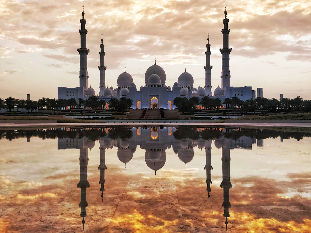 Reflection of the grand white mosque in Abu Dhabi at sunset, photo realistic, wide angle lens, shot on Sony Alpha A7 III with 24-35mm f/8 lens, sharp focus, beautiful reflections, clear sky, warm colors, golden hour, tranquil and peaceful atmosphere, serene desert landscape, golden sand and azure water, majestic architecture, serene mood in the style of a Sony Alpha A7 III photograph.