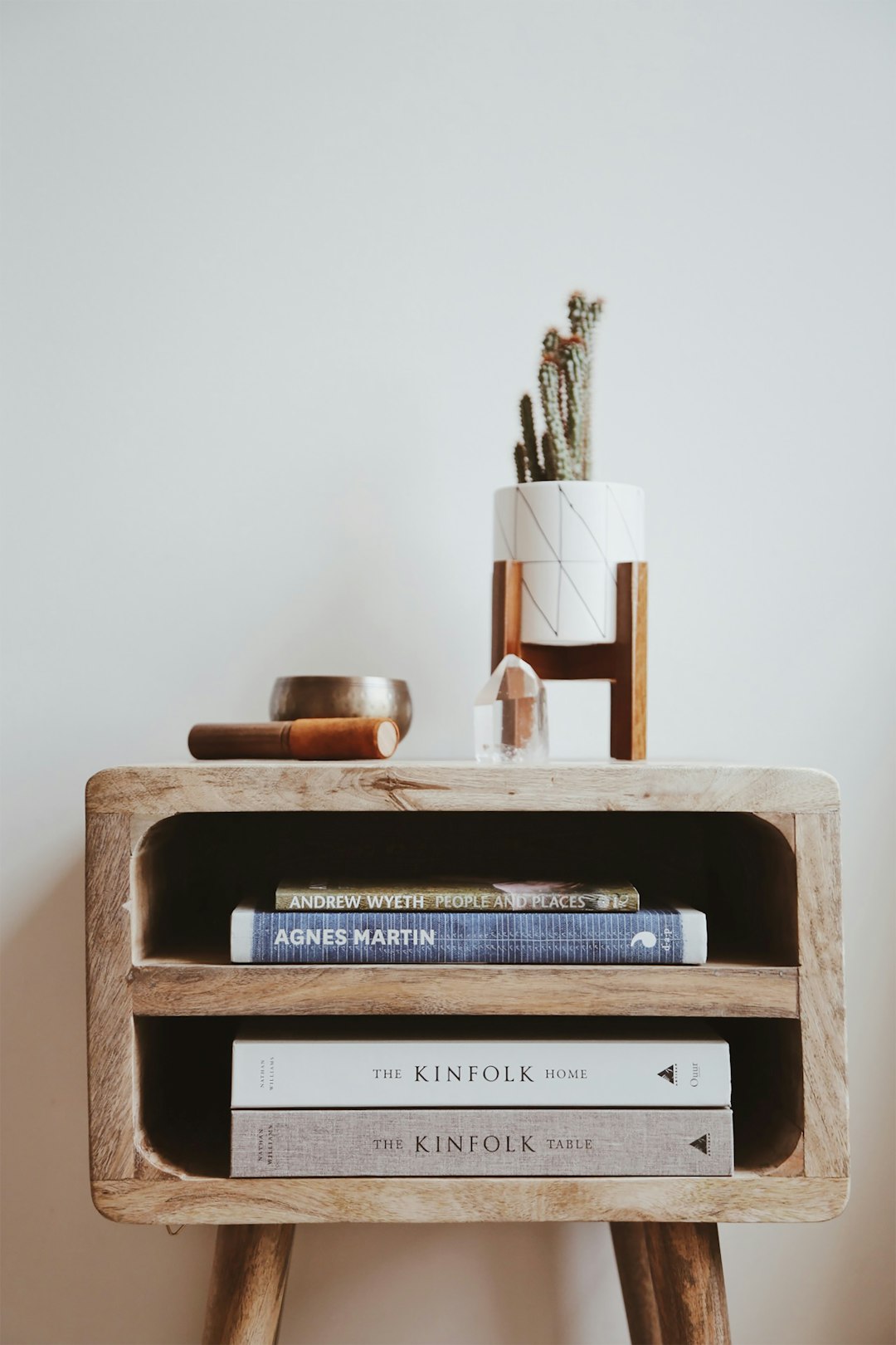 A wooden nightstand with books and cacti, a small glass jar on top of the table, white wall in the background, natural light, book titled “AND the wheel KIN Printed FOLDirty home The”. A minimalist design, simple yet elegant, focusing only on essential elements to convey clarity and simplicity in the style of minimalism. –ar 85:128