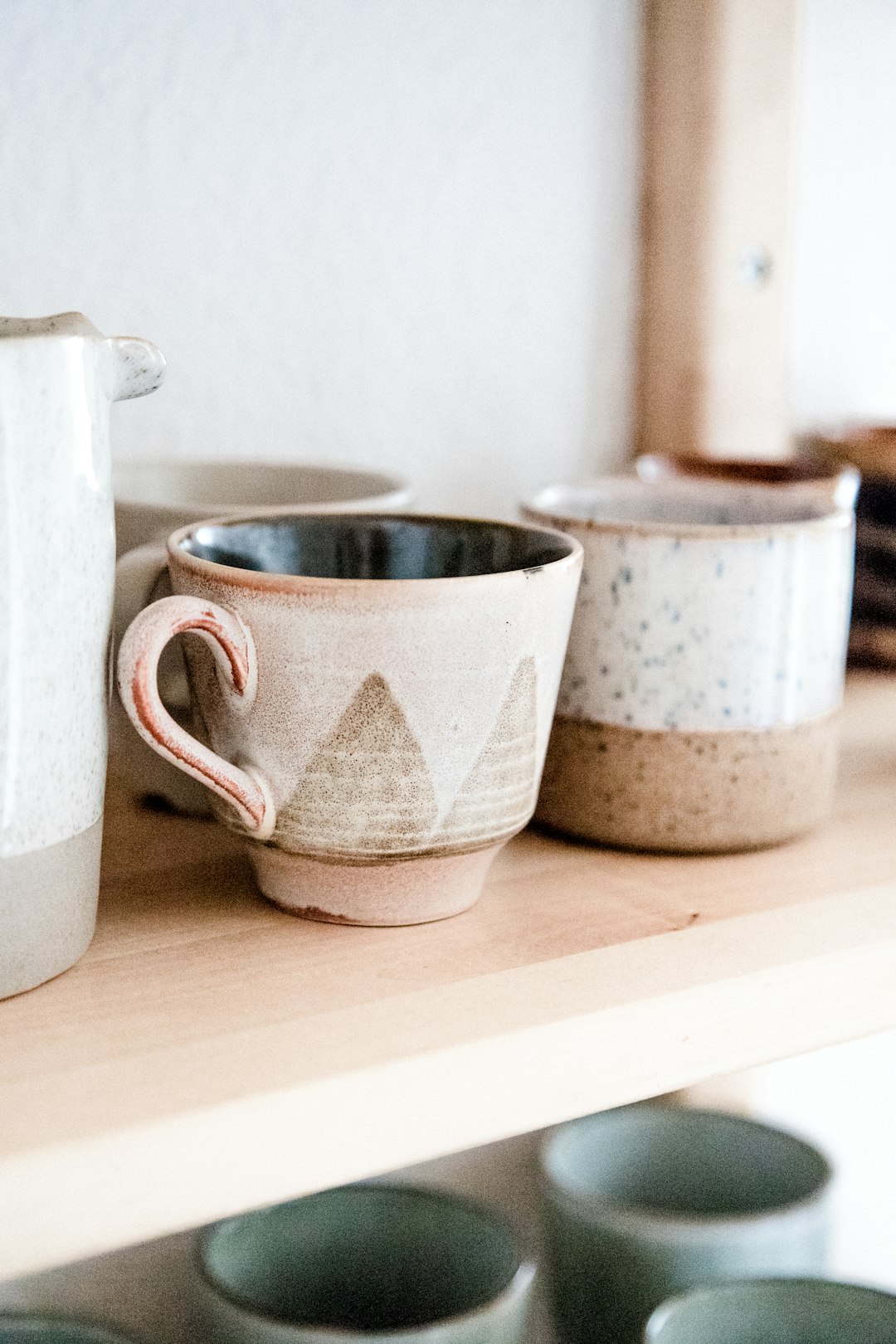 A closeup of handmade ceramic mugs and cups on a shelf, with a simple white wall in the background. The focus is on the unique shapes and textures of each mug, capturing their soft earthy tones against the bright light from a window, creating a cozy atmosphere that reflects home baking or handcrafted items. This shot would be perfect for product photography to highlight the individuality of the ceramics in the style of home baking or handcrafted items.