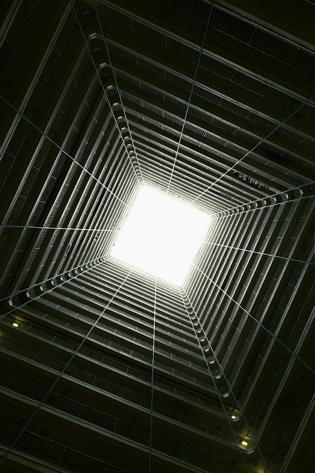 In the center of an apartment complex in Hong Kong, looking up at it from below through its skylight, you can see the high rise building’s gridlike architecture and a large square opening with sunlight shining down on it. The perspective is symmetrical, and there should be no other buildings around., photography, cinematic light –ar 85:128