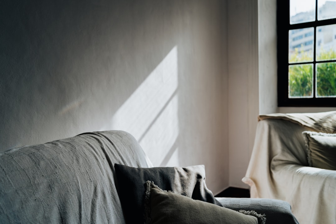 A sofa in an apartment with a window, a gray fabric cover on the couch, a window sill above it and sunlight shining through, a light colored wall behind the sofas, a blurred view of another living room outside, soft focus, shot in the style of Hasselblad X2D camera style raw. –ar 128:85